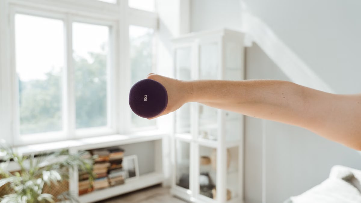 Close-up of a person lifting a 2kg dumbbell in a sunlit room.